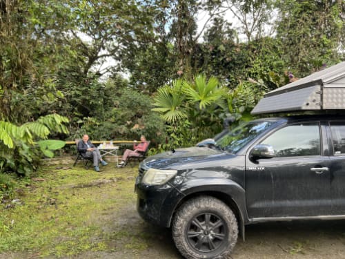 Two campers sit with their morning coffee with their camper van in the foreground.